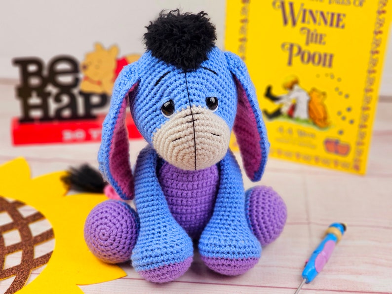 May include: A crocheted blue and purple Eeyore plush toy with a black yarn tail and a black yarn pom-pom on its head. The toy is sitting on a wooden surface with a yellow sunflower and a book titled "Winnie the Pooh" in the background.