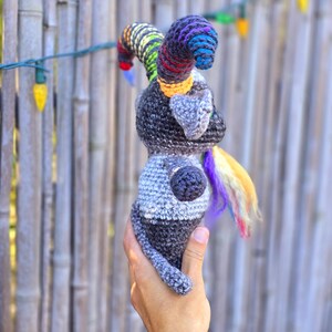 Puede incluir: Un animal de peluche gris tejido a crochet con un cuerno y una cola arco&iacute;ris. El animal est&aacute; siendo sostenido por una mano.