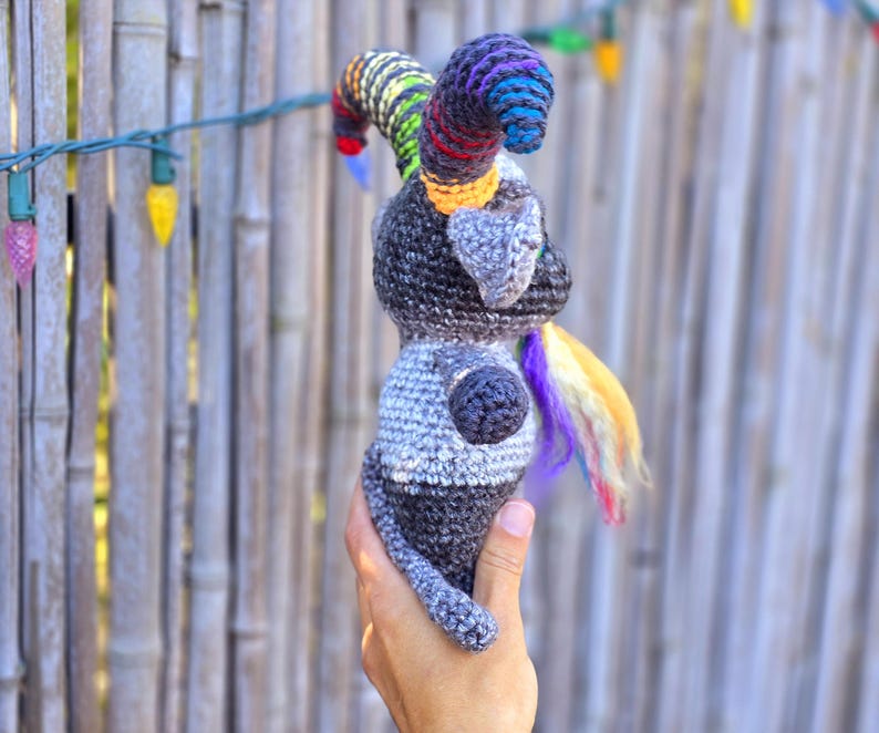 Puede incluir: Un animal de peluche gris tejido a crochet con un cuerno y una cola arco&iacute;ris. El animal est&aacute; siendo sostenido por una mano.