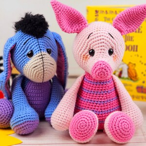 May include: Two crocheted stuffed animals, a blue donkey and a pink pig, sitting on a wooden surface. The donkey has a black mane and tail. The pig has a pink and purple striped shirt.