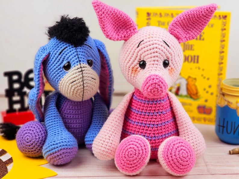 May include: Two crocheted stuffed animals, a blue donkey and a pink pig, sitting on a wooden surface. The donkey has a black mane and tail. The pig has a pink and purple striped shirt.