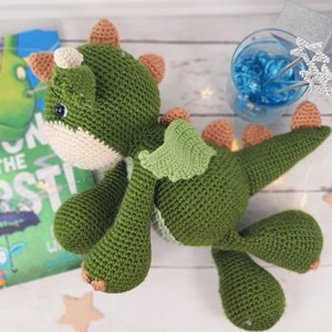 May include: A green crocheted dragon toy with a cream-coloured face and brown accents. The dragon has a green wing and is lying on a white surface.