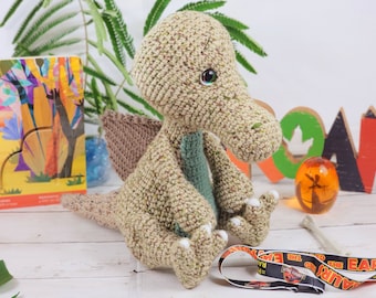 Pode incluir: Um brinquedo de dinossauro de crochê com um esquema de cores verde e marrom. O dinossauro tem uma cauda longa e um focinho pequeno e pontudo. Está sentado em uma superfície branca com um cordão do Jurassic Park em primeiro plano.