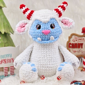 Pu&ograve; includere: Un giocattolo di peluche bianco lavorato a uncinetto con dettagli blu e rosa. Il giocattolo ha corna a strisce rosse e bianche e un viso blu con un sorriso pieno di denti. Il giocattolo &egrave; seduto su una superficie bianca con bastoncini di zucchero a strisce rosse e bianche e un cartello che dice "Kringle & Co. Candy Canes".