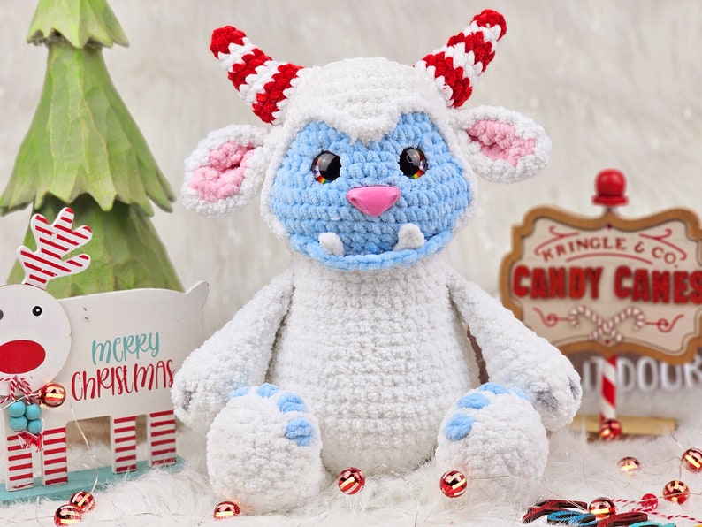 Pu&ograve; includere: Un giocattolo di peluche bianco lavorato a uncinetto con dettagli blu e rosa. Il giocattolo ha corna a strisce rosse e bianche e un viso blu con un sorriso pieno di denti. Il giocattolo &egrave; seduto su una superficie bianca con bastoncini di zucchero a strisce rosse e bianche e un cartello che dice "Kringle & Co. Candy Canes".
