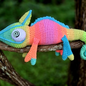 May include: A colorful, crocheted chameleon toy in shades of pink, orange, yellow, green, and blue, perched on a brown branch. The chameleon has a spiral eye design and is set against a blurred green background.