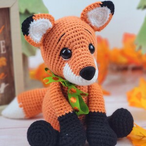 May include: A crocheted orange fox plush toy with black and white ears, black eyes, and a black nose. The fox is wearing a green ribbon with orange leaves around its neck. The fox is sitting on a white surface with fall leaves in the background.