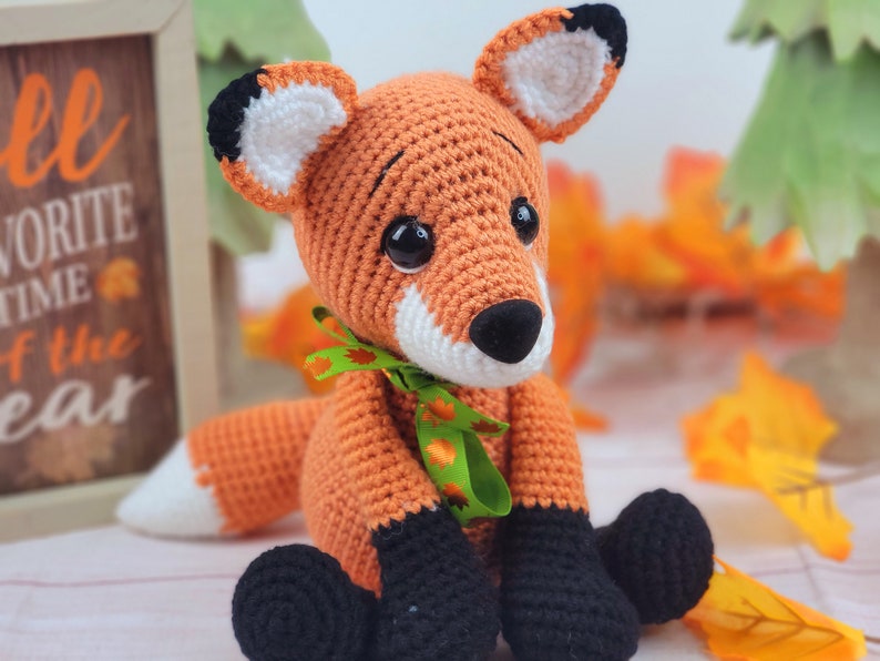 May include: A crocheted orange fox plush toy with black and white ears, black eyes, and a black nose. The fox is wearing a green ribbon with orange leaves around its neck. The fox is sitting on a white surface with fall leaves in the background.