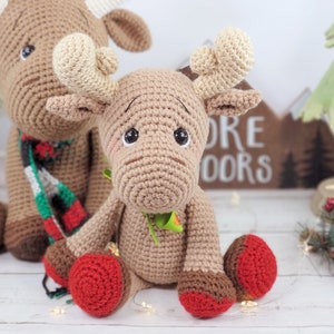 May include: Two crocheted moose toys, one brown with a red and green scarf, the other brown with green leaves around its neck. Both moose have red and brown hooves.