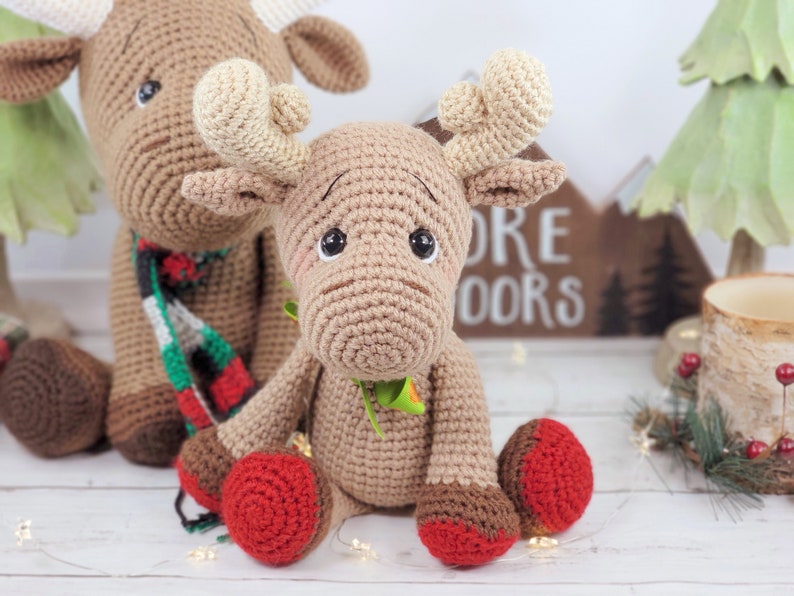 May include: Two crocheted moose toys, one brown with a red and green scarf, the other brown with green leaves around its neck. Both moose have red and brown hooves.