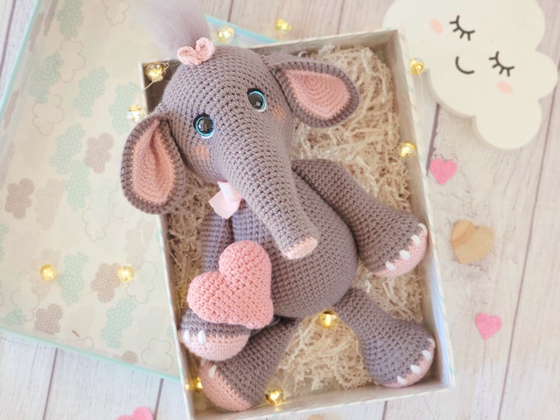 May include: A gray crocheted elephant holding a pink crocheted heart. The elephant has pink ears, a pink bow, and blue eyes. The elephant is sitting in a box with shredded paper.