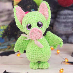 May include: A green crocheted bat with pink ears and a pink nose. The bat has large purple eyes and a pink ribbon around its neck.
