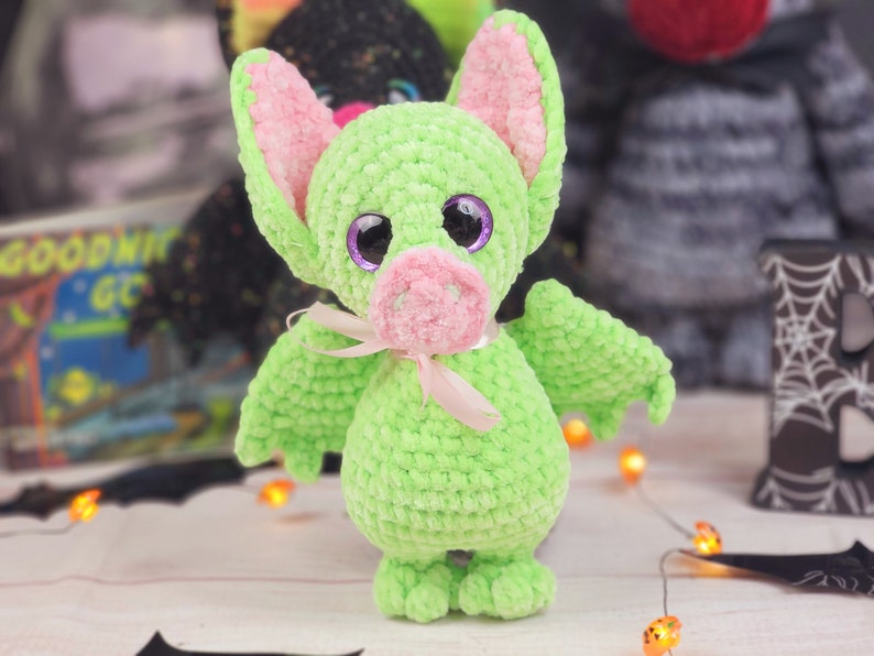 May include: A green crocheted bat with pink ears and a pink nose. The bat has large purple eyes and a pink ribbon around its neck.