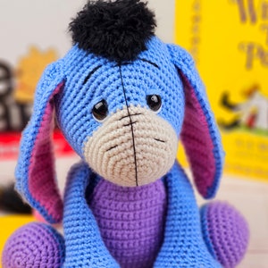 May include: A crocheted stuffed toy of Eeyore the donkey from Winnie the Pooh. The toy is blue and purple with a black yarn pom-pom on its head. It has a sad expression with black eyes and a stitched-on mouth.