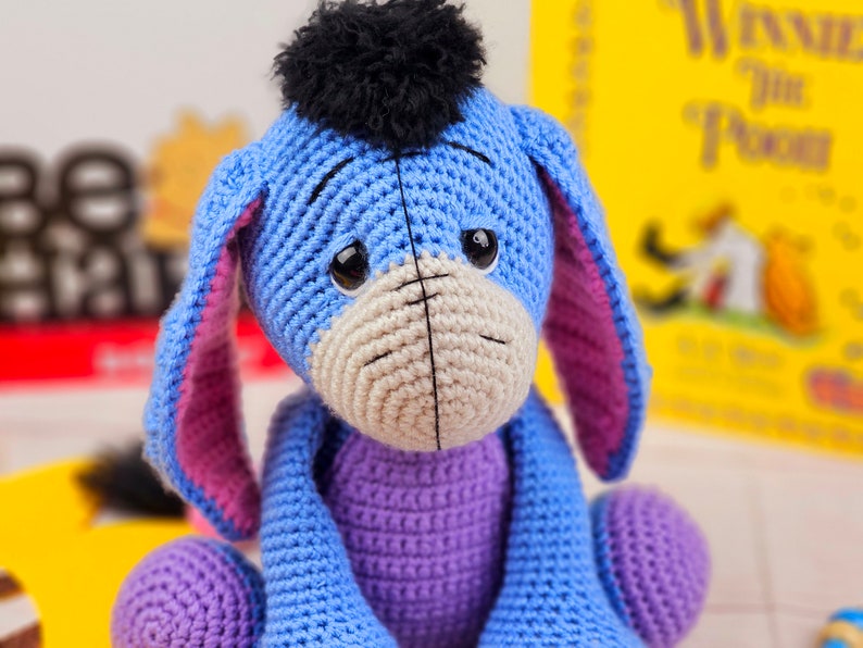 May include: A crocheted stuffed toy of Eeyore the donkey from Winnie the Pooh. The toy is blue and purple with a black yarn pom-pom on its head. It has a sad expression with black eyes and a stitched-on mouth.