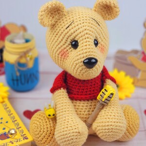K&ouml;nnte beinhalten: Ein geh&auml;keltes Winnie-the-Pooh-B&auml;ren-Spielzeug, das ein rotes Hemd tr&auml;gt und einen gelben und braunen Honigstock h&auml;lt. Der B&auml;r sitzt auf einem Tisch mit einem Buch und anderen Dekorationen.