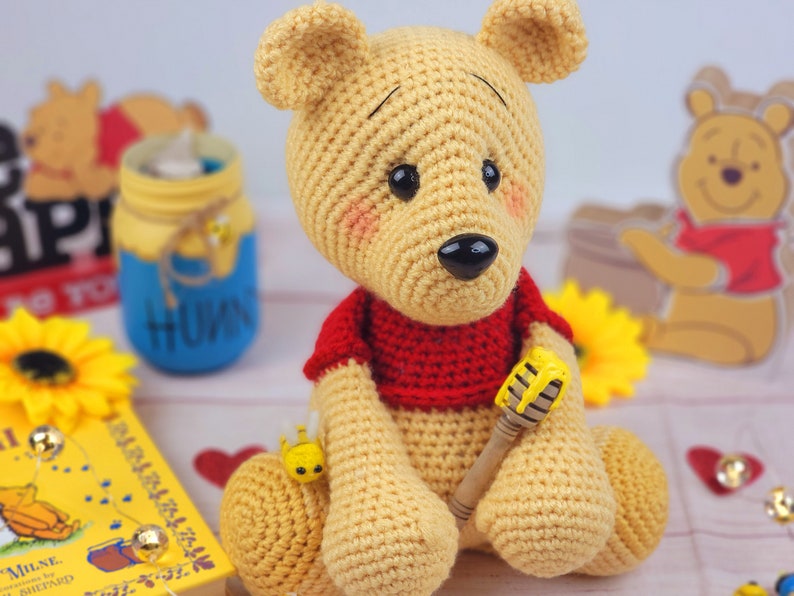 K&ouml;nnte beinhalten: Ein geh&auml;keltes Winnie-the-Pooh-B&auml;ren-Spielzeug, das ein rotes Hemd tr&auml;gt und einen gelben und braunen Honigstock h&auml;lt. Der B&auml;r sitzt auf einem Tisch mit einem Buch und anderen Dekorationen.