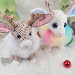 May include: Two crocheted reindeer dolls with large eyes and fluffy white fur. One is tan with pink-lined ears, the other is white with blue and pink-lined ears. Both have brown antlers and are sitting on a white surface with Christmas decorations.
