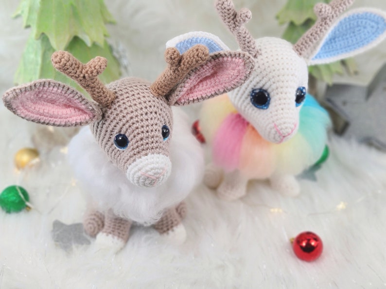 May include: Two crocheted reindeer dolls with large eyes and fluffy white fur. One is tan with pink-lined ears, the other is white with blue and pink-lined ears. Both have brown antlers and are sitting on a white surface with Christmas decorations.