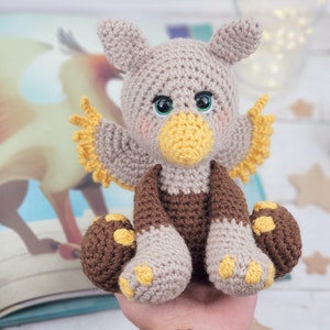 Pode incluir: Um animal de peluche de crochê em forma de grifo. O grifo é marrom, cinza e amarelo. Ele tem grandes olhos verdes e um bico amarelo. O grifo está sentado em uma mão.