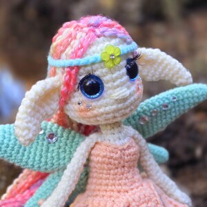May include: A crocheted fairy doll with pink and white hair, blue eyes, and green wings. The doll is wearing a peach-colored dress and a blue headband with a green flower.