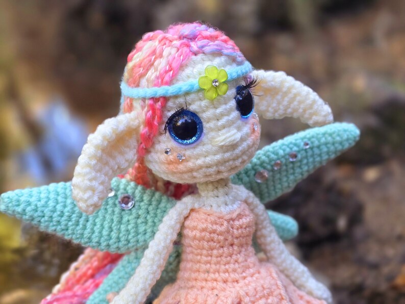 May include: A crocheted fairy doll with pink and white hair, blue eyes, and green wings. The doll is wearing a peach-colored dress and a blue headband with a green flower.