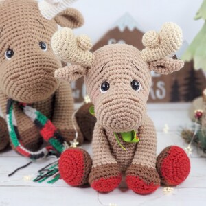 May include: Two crocheted moose toys, one brown with a red and green scarf and the other brown with green accents. Both moose have red and brown feet.