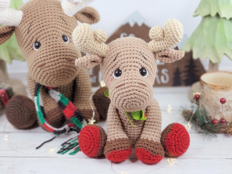 May include: Two crocheted moose toys, one brown with a red and green scarf and the other brown with green accents. Both moose have red and brown feet.