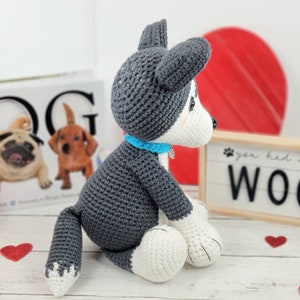 May include: A crocheted gray and white husky dog plush toy sitting on a white surface. The dog is wearing a blue collar and has a white patch on its chest. The dog is looking to the right. The background includes a book with the word "DOG" on the cover, a red heart, and a sign that says "You had me at woof."