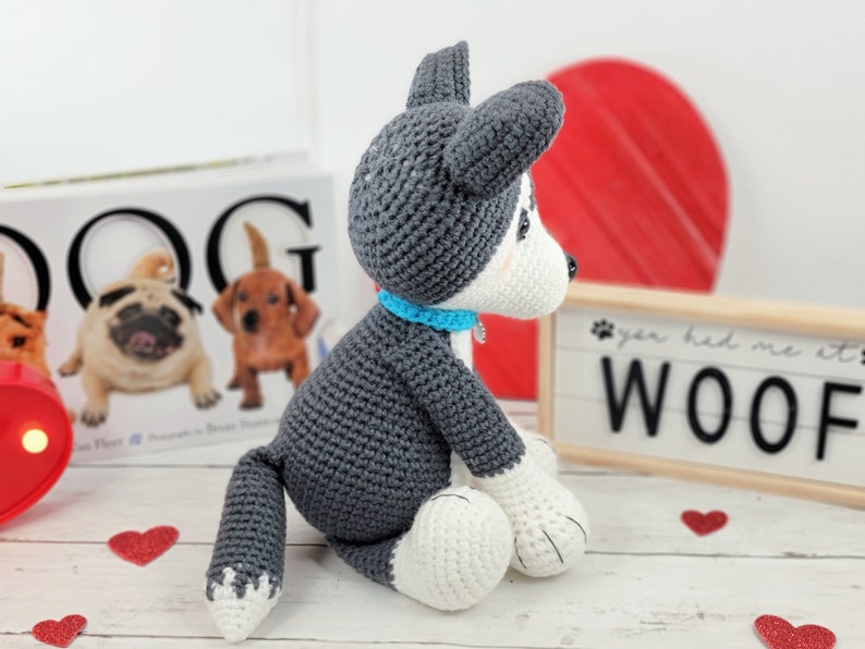 May include: A crocheted gray and white husky dog plush toy sitting on a white surface. The dog is wearing a blue collar and has a white patch on its chest. The dog is looking to the right. The background includes a book with the word "DOG" on the cover, a red heart, and a sign that says "You had me at woof."