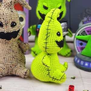 Puede incluir: Dos juguetes de peluche de crochet, uno verde y otro marr&oacute;n claro, est&aacute;n sentados sobre una superficie de madera blanca. Ambos juguetes tienen forma de fantasmas con ojos y bocas cosidos. El fantasma verde tiene un bigote negro cosido.