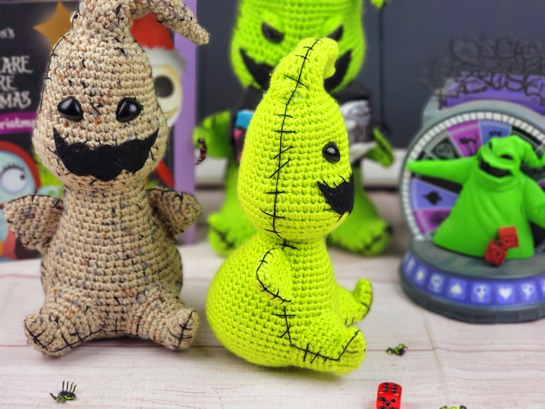 Puede incluir: Dos juguetes de peluche de crochet, uno verde y otro marr&oacute;n claro, est&aacute;n sentados sobre una superficie de madera blanca. Ambos juguetes tienen forma de fantasmas con ojos y bocas cosidos. El fantasma verde tiene un bigote negro cosido.