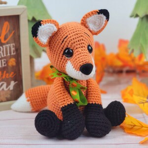 May include: A crocheted orange fox with black feet and a green ribbon around its neck. The fox is sitting on a white wooden surface with fall leaves scattered around it.