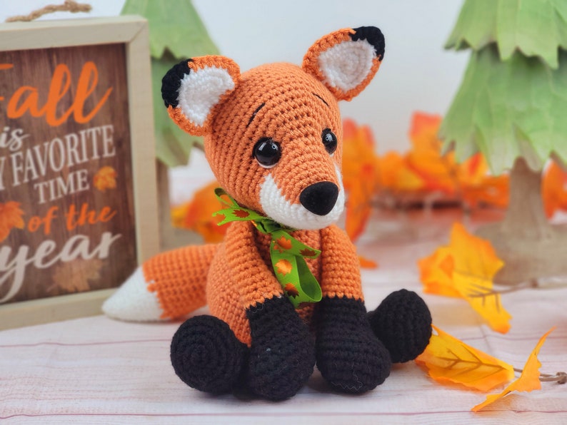 May include: A crocheted orange fox with black feet and a green ribbon around its neck. The fox is sitting on a white wooden surface with fall leaves scattered around it.