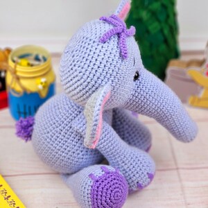 May include: A crocheted purple elephant toy with a pink and white ear, a purple flower on its head, and black eyes. The elephant is sitting on a white wooden surface.
