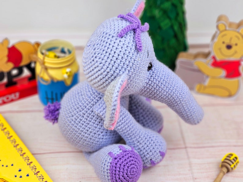 May include: A crocheted purple elephant toy with a pink and white ear, a purple flower on its head, and black eyes. The elephant is sitting on a white wooden surface.