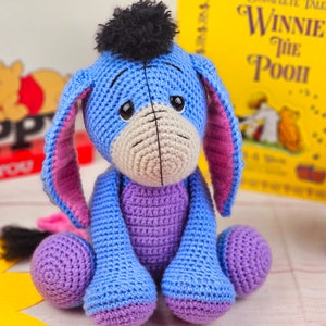 May include: A crocheted blue and purple Eeyore plush toy with a black pom-pom on its head. The toy is sitting on a wooden surface with a book titled "Winnie the Pooh" in the background.