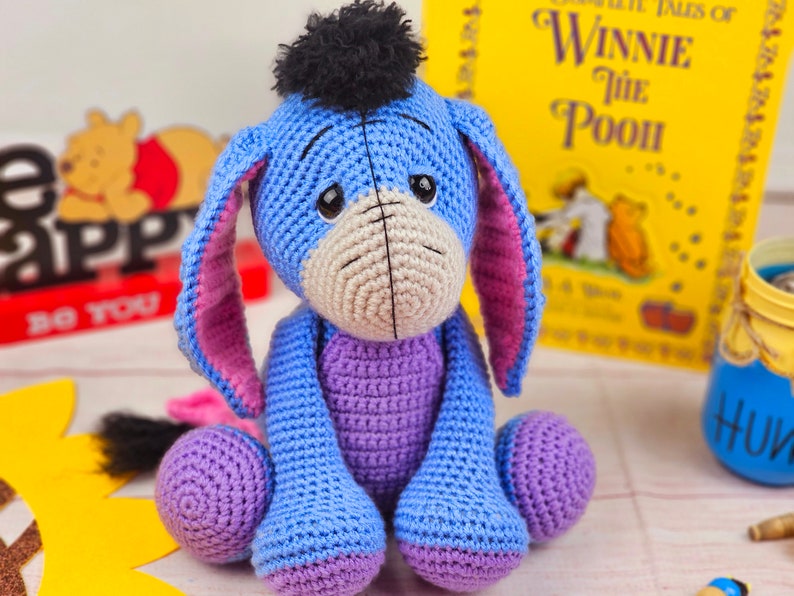 May include: A crocheted blue and purple Eeyore plush toy with a black pom-pom on its head. The toy is sitting on a wooden surface with a book titled "Winnie the Pooh" in the background.