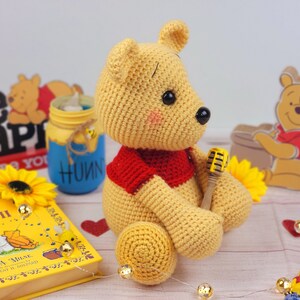 K&ouml;nnte beinhalten: Ein geh&auml;kelter Winnie Puuh B&auml;r, der ein rotes Hemd tr&auml;gt und ein gelbes Mikrofon h&auml;lt. Der B&auml;r sitzt auf einer rosa Oberfl&auml;che mit einem Buch, einem Glas und einer Sonnenblume.