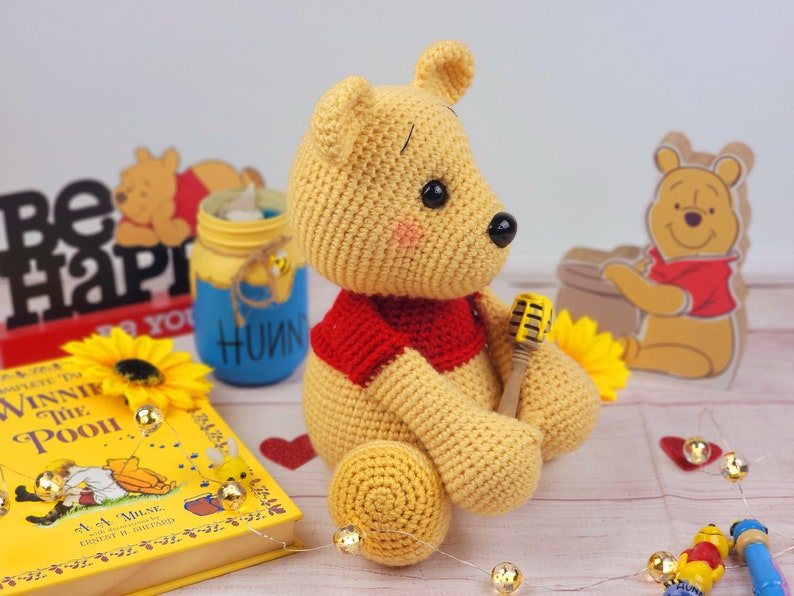 K&ouml;nnte beinhalten: Ein geh&auml;kelter Winnie Puuh B&auml;r, der ein rotes Hemd tr&auml;gt und ein gelbes Mikrofon h&auml;lt. Der B&auml;r sitzt auf einer rosa Oberfl&auml;che mit einem Buch, einem Glas und einer Sonnenblume.