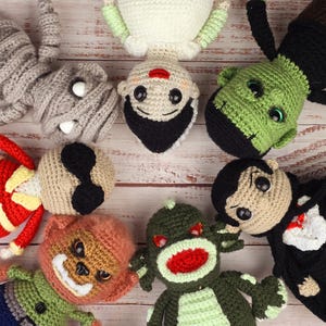 Puede incluir: Un grupo de peluches de monstruos de crochet, incluyendo una momie, una novia, Frankenstein, Drácula, un mono, un dragón y un zombie. Los peluches están dispuestos en un círculo sobre un fondo de madera blanco.