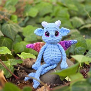 Puede incluir: Un pequeño dragón azul de crochet con alas rosas y moradas se sienta en una roca en un lecho de hojas verdes.
