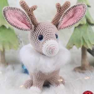 May include: A crocheted reindeer with a light brown body, pink-lined ears, and blue eyes. It has a white fluffy collar and small antlers. The reindeer is a handmade Christmas decoration.