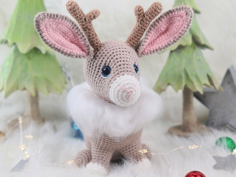 May include: A crocheted reindeer with a light brown body, pink-lined ears, and blue eyes. It has a white fluffy collar and small antlers. The reindeer is a handmade Christmas decoration.