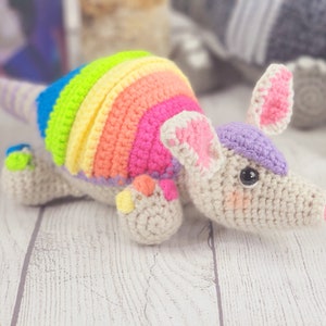 May include: A crocheted armadillo toy with a rainbow shell and a purple and pink head. The toy is made with soft yarn and has a sweet expression.