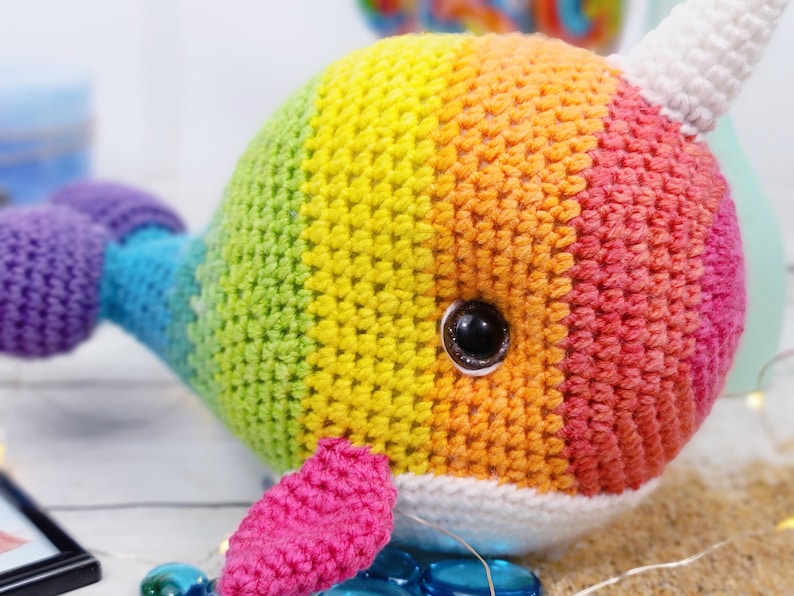 K&ouml;nnte beinhalten: Ein regenbogenfarbenes, geh&auml;keltes Narwal-Pl&uuml;schtier mit einem wei&szlig;en Horn und einem schwarzen Auge. Das Spielzeug ist aus verschiedenen Farben gefertigt, darunter Lila, Blau, Gr&uuml;n, Gelb, Orange und Rot.