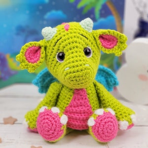 Puede incluir: Un drag&oacute;n de crochet verde con orejas, alas y pies rosas. El drag&oacute;n tiene ojos grandes y negros y una barriga rosa.