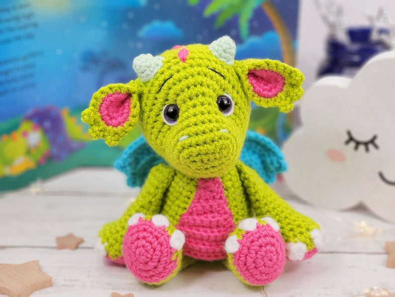 Puede incluir: Un drag&oacute;n de crochet verde con orejas, alas y pies rosas. El drag&oacute;n tiene ojos grandes y negros y una barriga rosa.
