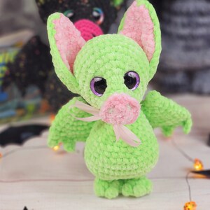 May include: A green crocheted bat with pink ears and a pink nose. The bat has large purple eyes and a pink ribbon around its neck.