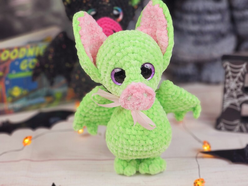 May include: A green crocheted bat with pink ears and a pink nose. The bat has large purple eyes and a pink ribbon around its neck.